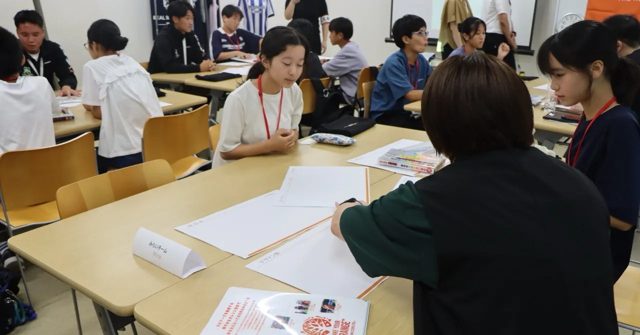 グループの机を囲み、アドバイザーの社員と話しながら企画書を書く生徒たち