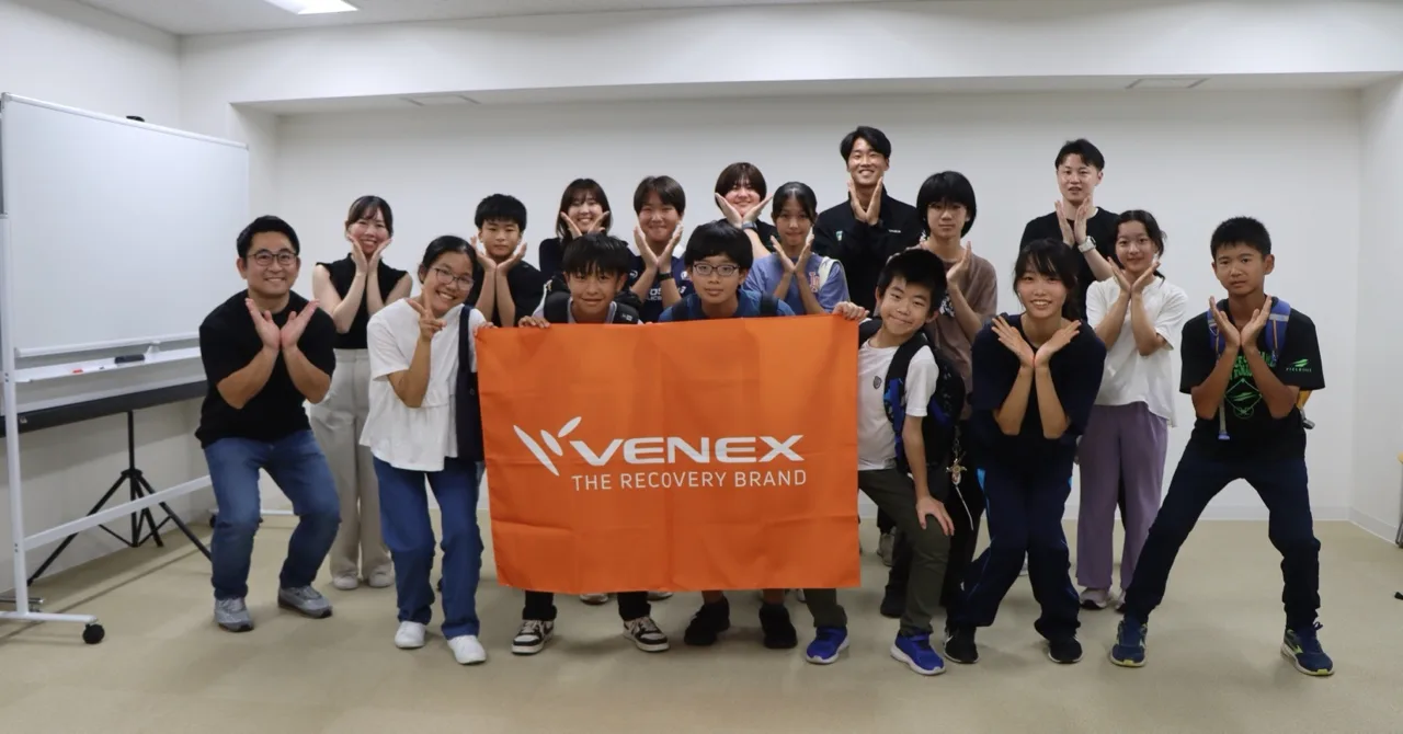VENEXのフラッグを囲み、Vマークのポーズで並ぶ参加者全員の集合写真