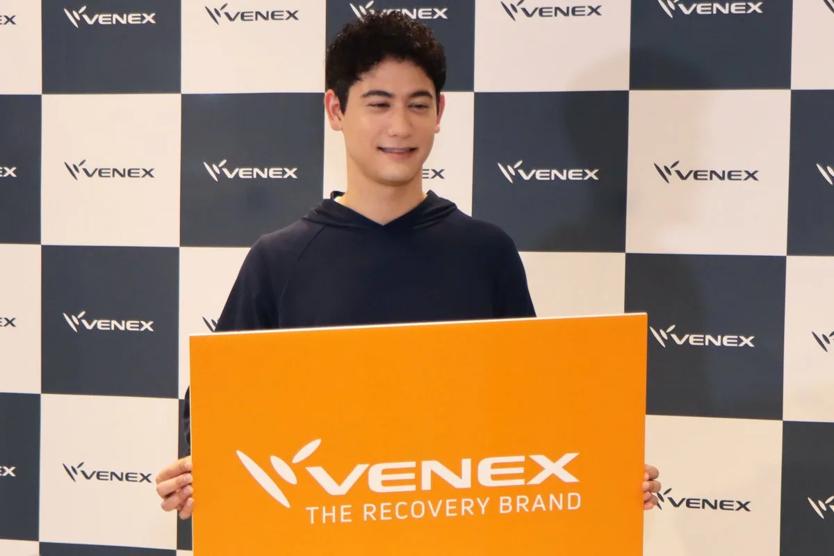 VENEXのロゴパネルを手に持ち微笑む国山ハセンさん（ネイビーのフーディを着用）