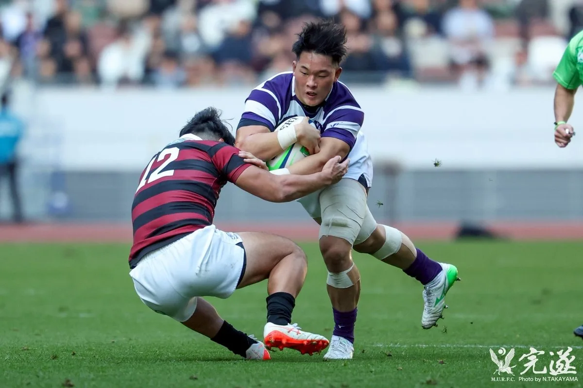 試合中、相手選手のタックルを受けながら前進する明治大学の選手