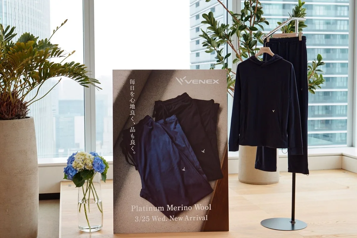 会場に置かれた「Platinum Merino Wool 3/25 New Arrival」のパネルと、ラックに掛かったネイビーのフーディとパンツ